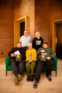 David, Louise and Dave sit on a sofa holding their awards. Two people stand behind them. 